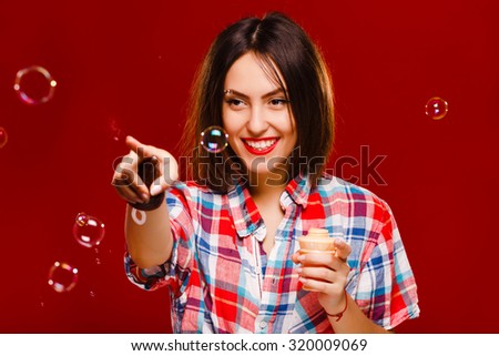 Cheerful girl, with dark hair, wearing in red checkered shirt,  playing with bubbles, on red background, in studio, waist up