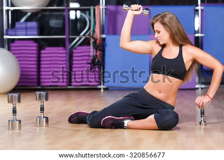 Fitness woman, with long hair, wearing in sneakers black top and pants, does exercises with dumbbells on the floor, in the gym, full body
