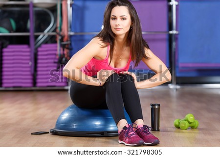 Pretty fitness woman, wearing in shirt, sneakers and black leggings, sitting on a bosu ball and looking at camera, cup, dumbbells and smart phone lying on the floor, in the gym, full body
