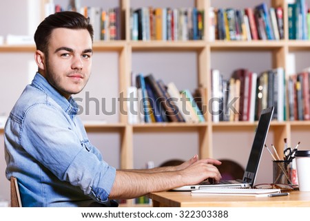 Side view of a handsome man, wearing in casual blue shirt, working on laptop and looking at camera, in a bright office, waist up