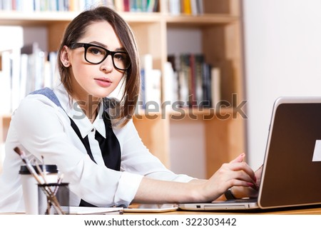 Beautiful brunette woman, wearing in white and black blouse and glasses, sitting at the table with laptop and looking at camera, on the bookshelves background, in office, waist up