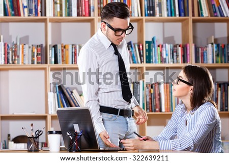 Young confused man, wearing in white shirt, glasses and tie, standing near beautiful woman\'s table and they are talking about work, in modern office on the bookshelves background, waist up