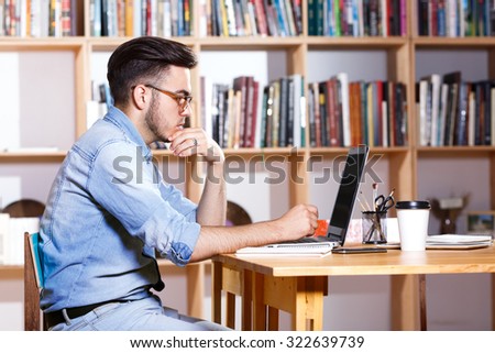 Side view portrait of serious man, wearing in casual shirt and glasses, sitting at the table and looking at the laptops screen, on the bookshelves background, waist up