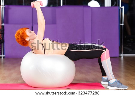 Beautiful fitness girl with short haircut wearing in top and leggings does exercises with dumbbells and white fitball on a pink yoga mat, in the gym, full body