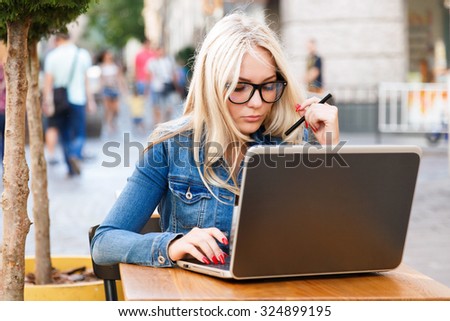 Pretty young woman, wearing in glasses, white shirt and blue jacket, sitting at the table and working with laptop, on the street of old city on summer day, waist up