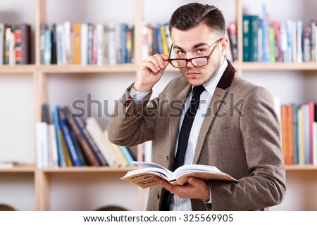 Serious young man, wearing in glasses white shirt and jacket, posing with a book, on the bookshelves background, in the office, waist up
