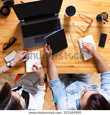 Point of view of young business woman and man, wearing in casual clothes, sitting at the table with notebooks, tablet and laptop, in the office, waist up
