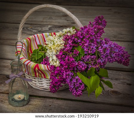 Basket with a bouquet of lilacs and light with a burning candle in the evening on a wooden table