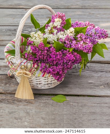 Bouquet of lilac in drops of dew in a white basket and straw angel figurine on wooden table