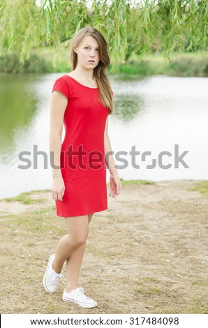 Young beautiful girl with flowing hair and freckles on her face in a red dress and white sneakers standing in the park. Green background and the lake