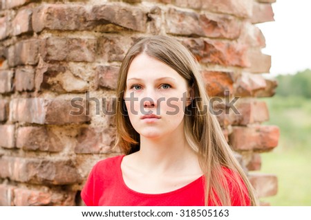 The young beautiful girl with freckles and red dress standing near the wall of red brick