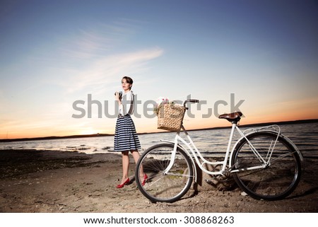 Sunset at the seaside. Younggirl in a lovely dress is posing near the white cycle.