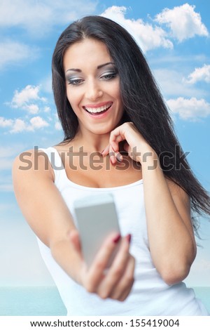Portrait of happy young woman taking photos with mobile phone camera on sky background