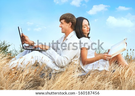 Beautiful couple sitting on grass with laptop and book against sky background