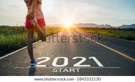 Photo of New year 2021 or start straight concept.word 2021 written on the asphalt road and athlete woman runner stretching leg preparing for new year at sunset.Concept of challenge or career path and change.