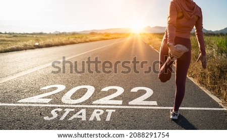New year 2022 or start straight concept.word 2022 written on the asphalt road and athlete woman runner stretching leg preparing for new year at sunset.Concept of challenge or career path and change.
