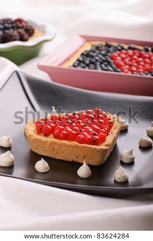Heart-shaped tart with redcurrants served on a black plate decorated with small meringues. Tarts with berries out of focus in the background. Selective focus, shallow DOF