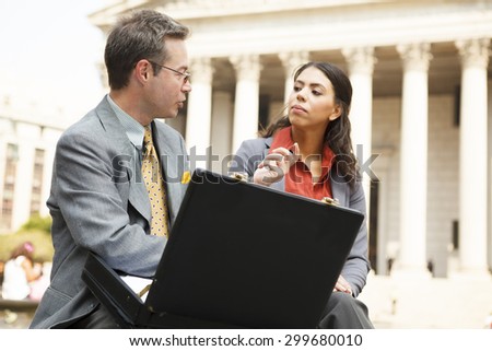 A seated man with an open briefcase talking to a woman as she listens attentively. A stately courthouse type building with columns is in the background. Focus is on the man.