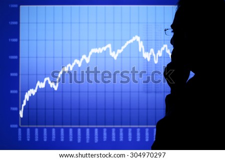 A man in silhouette is contemplating a rising financial or stock chart displayed on an LCD screen. Focus is on the man\'s eyeglasses. Chart created by the photographer.