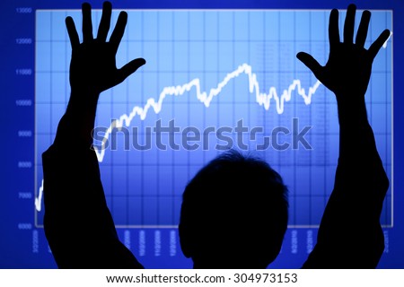 Silhouette of a man with his hands raised up in front of a rising stock chart on an LCD display. Focus is on man\'s hands and arms. Chart created by the photographer.