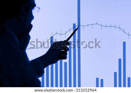 Silhouette of a man pointing to a rising graph on an LCD display with a mechanical pencil. Focus is on the LCD display.