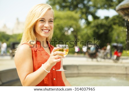 A smiling young woman holds a  glass of wine as she stands by a park fountain.
