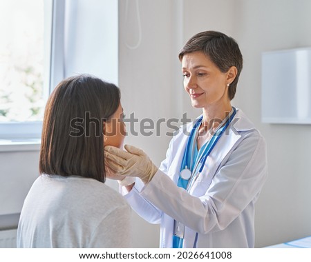 Friendly middle-aged woman doctor wearing gloves checking sore throat or thyroid glands, touching neck of young African female patient visiting clinic office. Thyroid cancer prevention concept ストックフォト © 