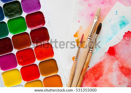 Palette of watercolor paints, brushes and paper for a water color on white background, close up.