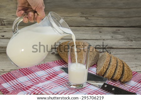 hand pouring milk from a jug into a glass on a wooden background grain bread on a napkin and knife