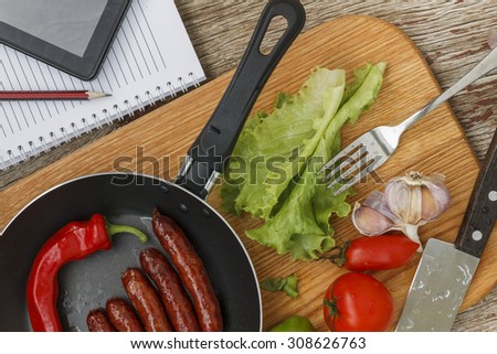 healthy Eating. frying pan with roasted Bavarian sausages and chili hot pepper on wooden background with vegetables and tablet PC, notebook and pen. business lunch