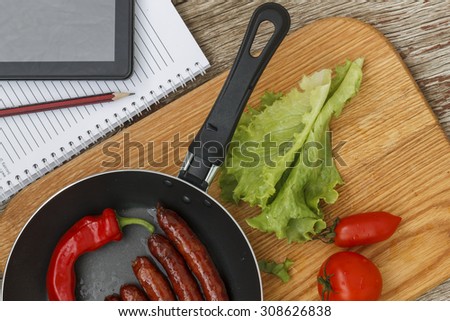healthy Eating. frying pan with roasted Bavarian sausages and chili hot pepper on wooden background with vegetables and tablet PC, notebook and pencil. business lunch