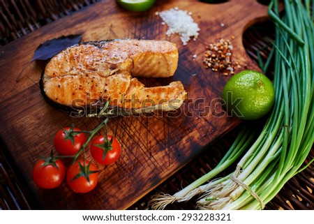 ?ook restaurant cooked a salmon steak on the grill and served with him a piece of lemon, onion, few tomatoes. Fish lying on a wooden surface, close to her small tomatoes on a branch, onion and lime
