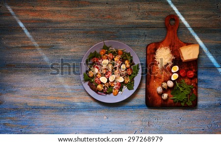 On blue wooden table is a plate with a light salad of tomato, eggs, greens and parmesan salad dressed with olive oil and balsamic vinegar, standing next to the board which was shredded ingredients