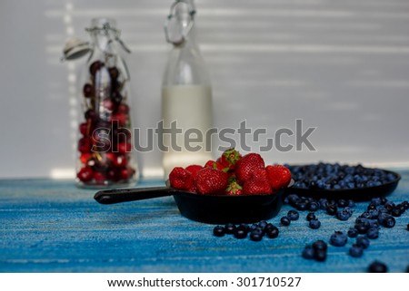 On a blue vintage wooden desk are two cast iron pans that sprinkling strawberries and blueberries