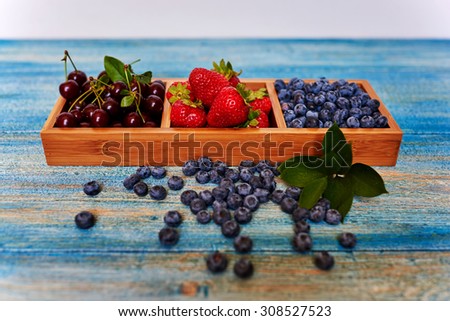 On the table lies a blue vintage wooden form with individual cells for a variety of sweets and fruits, cook it spread fresh ripe blueberries, strawberries and cherries