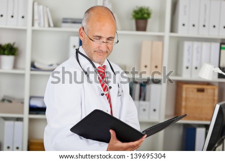 Mature male doctor reading folder against shelves in office