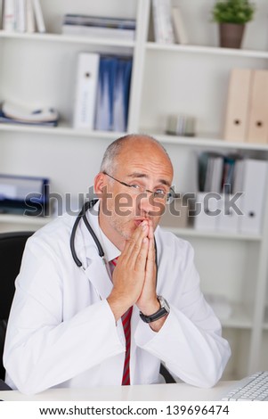 Portrait of thoughtful mature doctor with hands clasped sitting at office desk