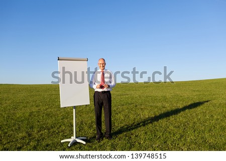 Portrait of mature businessman standing by blank flipchart on grassy field against clear sky
