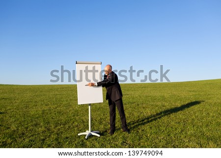 Mature businessman with pointing at flipchart on grassy field against clear sky