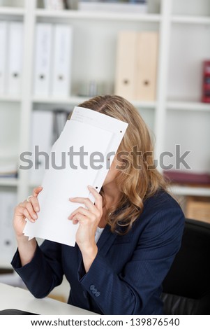Irritated businesswoman covering face with documents at office desk