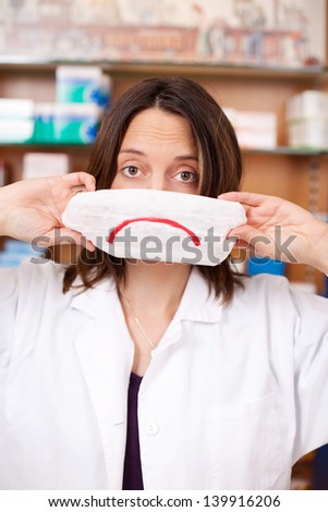 Portrait of mid adult female pharmacist holding mask with sad smiley in pharmacy