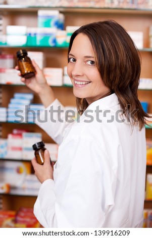 Portrait of happy female pharmacist holding medicine bottles while looking over shoulder against shelves in pharmacy
