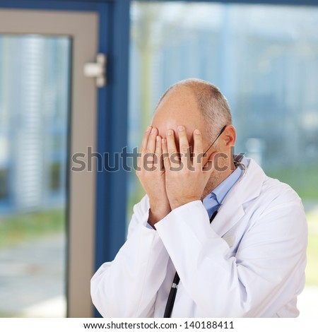 Mature upset doctor covering face with hands in clinic