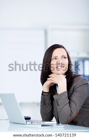 Pensive young business woman sitting at her desk in the office with her laptop in front of her reminiscing or planning a business strategy