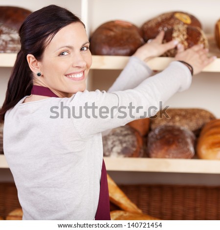 Friendly female bakery employee looking back over her shoulder and smiling as she reaches for a loaf bread on a shelf