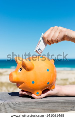 Closeup of woman\'s hand inserting euro note in piggy bank at beach
