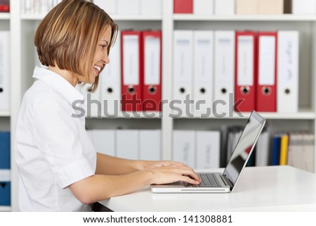 Side view of smiling businesswoman sitting in office with laptop computer, looking at computer