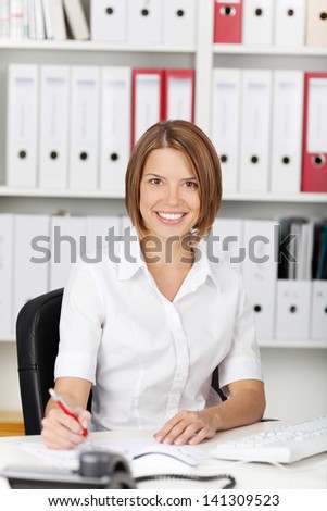 Smiling woman sitting at her desk in the office with a backdrop of shelves stacked with files