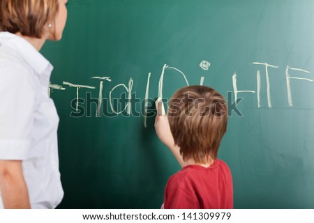 Young boy writing on the blackboard with chalk watched by his teacher