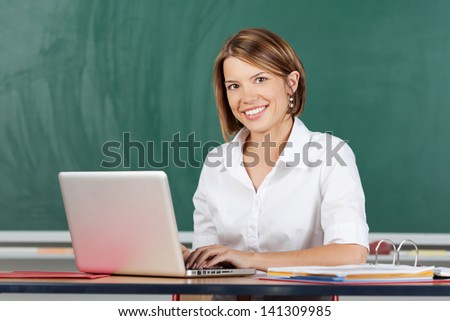 Happy woman typing on a laptop sitting at a table in front of green chalkboard smiling at the camera